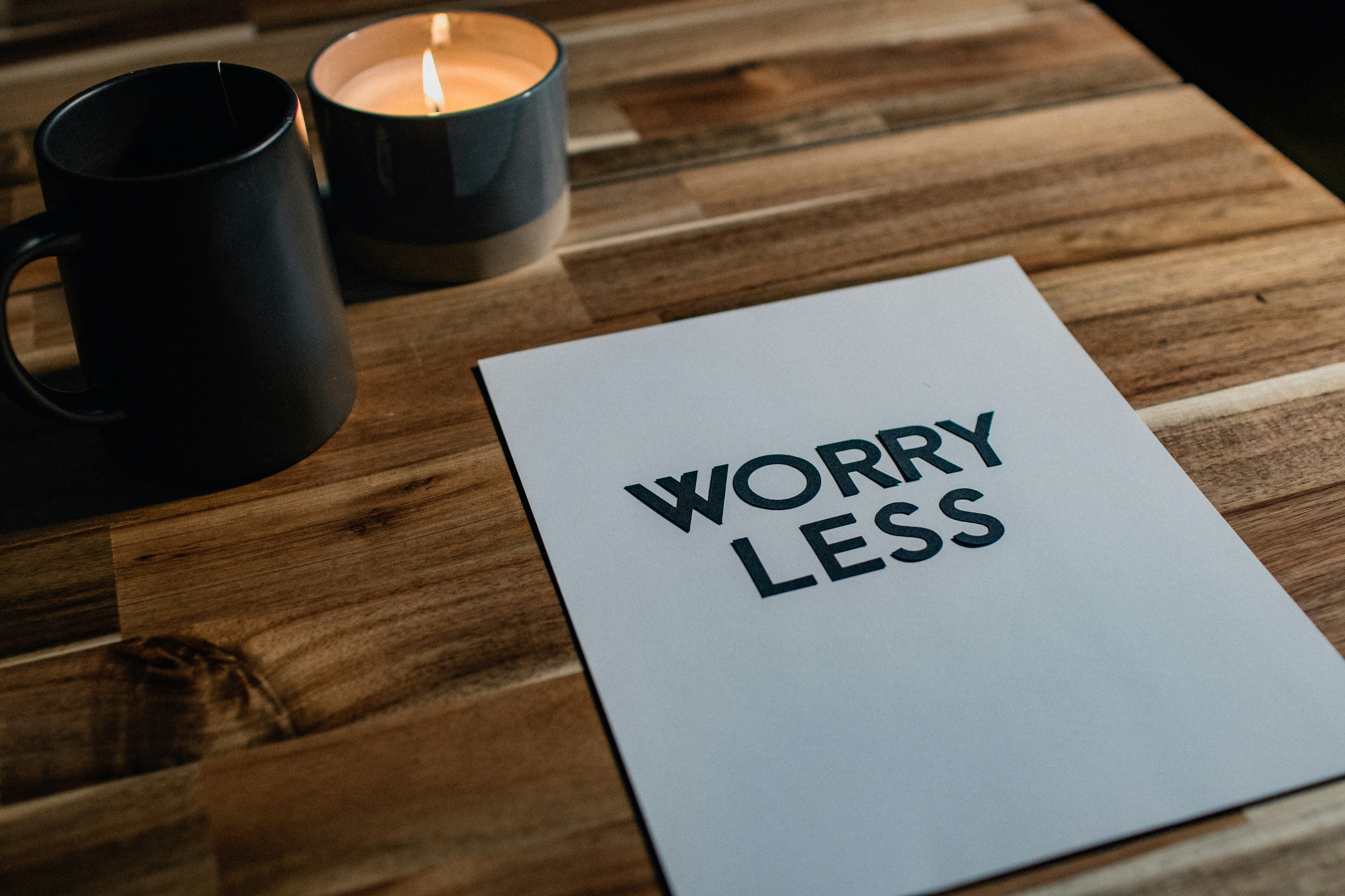 Eine brennende Kerze, eine Tasse und eine Karte mit der Aufschrift „WORRY LESS“ auf einem Holztisch. Stressfreie Routine.