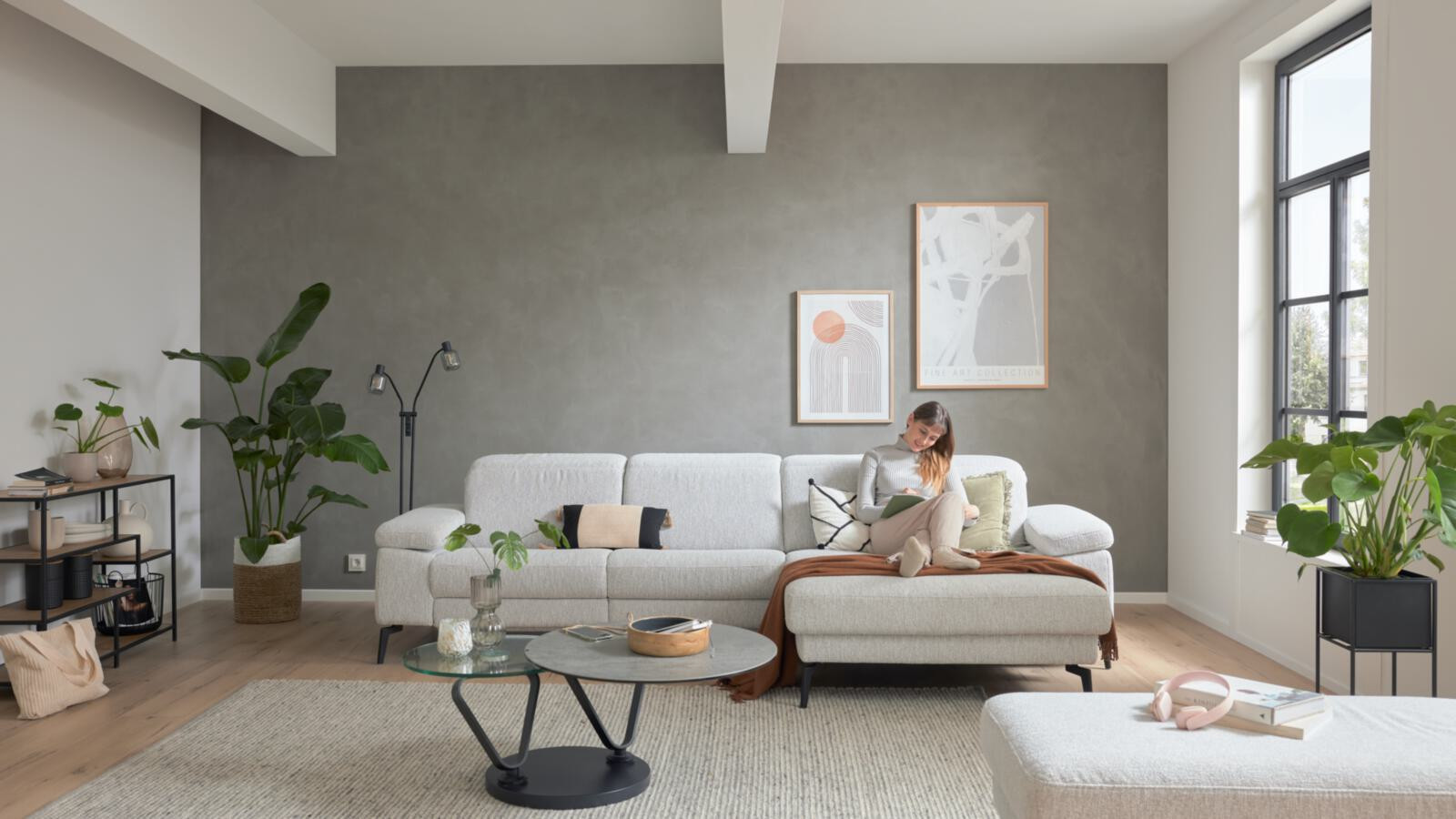 Frau liest auf einem hellen Sofa in einem modernen Wohnzimmer mit Pflanzen und Kunstwerken.